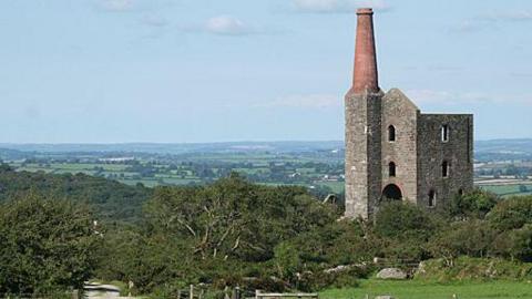 A rural landscape shows an old stone engine house with a tall chimney standing on a hillside beside a winding dirt path, fields, and low stone walls stretching across the countryside.