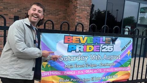 A man - Daniel Rothery - stood in front of a black fence pointing with both hands to a banner hanging from the fence. He is smiling at the camera, wearing a light grey jacket and black trousers. He has a brown moustache and a short brown fringe with a mullet. The banner is bright and multi-coloured. It reads: "BEVERLEY PRIDE 25. Saturday 16th August. Main Event 12-6pm - Flemingate. LIVE MUSIC - DANCE - DJS - DRAG ACTS - STALLS - WORKSHOPS. Pride Part 6pm til late at FIREPIT".