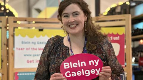Ruth Keggin-Gell, who has long wavy brown hair, is wearing a dark-coloured patterned top and holding a red card shaped like a speech bubble that says Blein ny Gaelgey.
