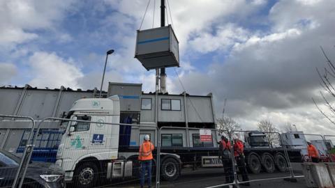 A crane with a white cab is using chains to grab a grey metal cabin above a complex of grey cabins, arranged in two stories. Three people in orange hi-vis jackets and white hard hats are looking upwards to watch the movement. There is metal fencing in the foreground. The sky is cloudy.