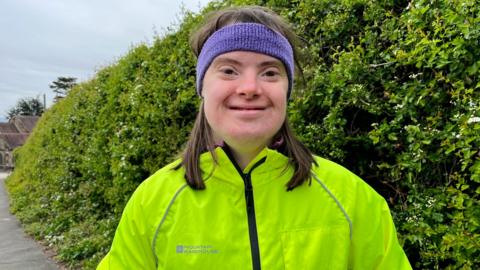 A woman in a lime green running jacket and a purple headband stood in front of a green hedge