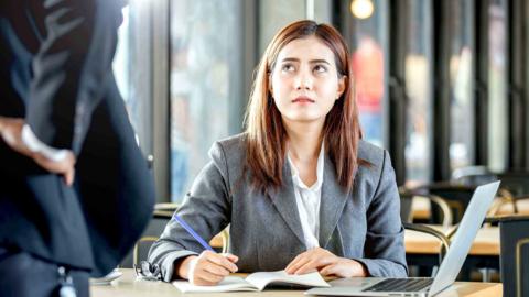 Young woman sat at work, looks up at someone wearing a suit with a hand on their hip