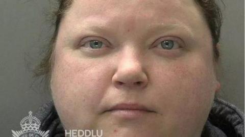 Jemma Lewis has blue eyes and brown hair. She is wearing a top with a grey collar. The top of the Gwent Police logo can be seen in this police mugshot.