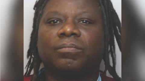 A police mugshot of William Kyaruhanga. He had dreadlocks and is standing against a pale white backdrop.