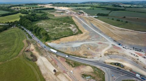 A bird's eye view of part of the A417 construction site stretching off into the distance with an active road to the left. The roads are surrounded by fields and trees.