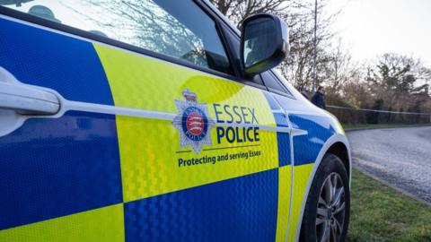 A blue and yellow police car parked on the side of a rural road. Essex Police is written on its door, with the Essex Police crest. 