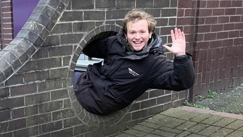 A man in a black coat, waving at the camera as he goes through a round hole in a brick wall