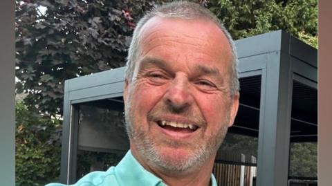 A selfie of Mark Beech who is smiling and has grey hair and facial hair. He is wearing a bright teal blue shirt and is in a garden with trees in the background