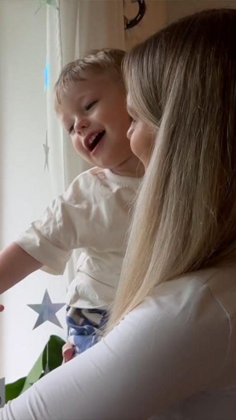 A young woman stands looking out the window holding a blonde haired boy.