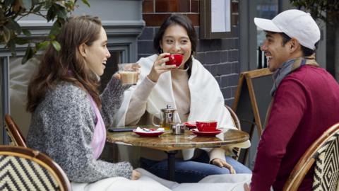 Two women and a man sit at an outdoor table drinking coffee.