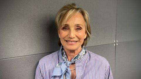 Kristin Scott Thomas, wearing a purple striped shirt and a blue neckerchief, standing in front of a grey wall