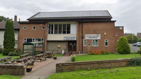 Exterior of the club. It is a two-storey brown brick building and has large windows and solar panels on the roof. There are wooden tables and chairs on a grassed area in front of the club alongside a brick path.