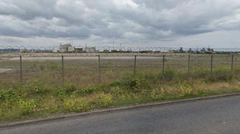 An empty industrial plot at Seal Sands near Billingham. A metal fence topped with barbed wire is by the roadside and scrubland is beyond the fence. It is an overcast day with a thick bed of clouds in the sky.