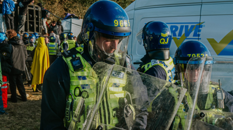 Three police officers in riot gear stand near other officers, rave attendees and a van