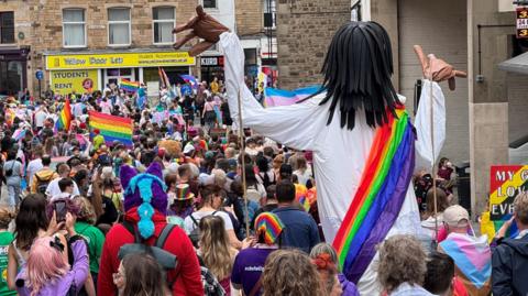 A large marionette puppet in a white robe and rainbow sash towers above a crowd of people walking through the city centre. Shops can be seen in the background