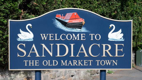 A welcome to Sandiacre road sign which has two swans and a canal boat featured on it