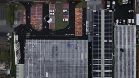 Three buildings covered in solar panels can be seen, with parking spaces and grass to the side
