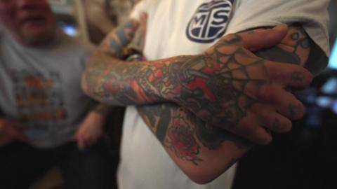 A close up of a tattooed arm. The person is wearing a white t-shirt. Their tattoo uses red and green ink. 
