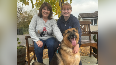  Cathy Shilling and daughter Katie, pictured with dog Max, who appears to be a German Shephard. The two women are sitting on a bench. Cathy is wearing a grey Christmas themed jumped and Katie is in an RSPCA fleece