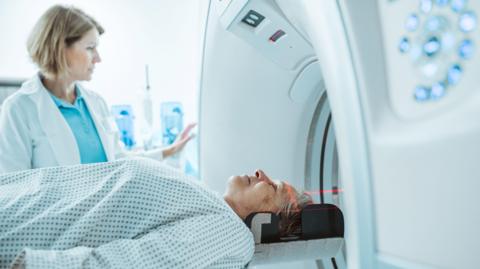 An older woman with light fair hair and wearing a hospital gown lies down at the entrance to a large circular MRI scanner. Standing next to her is a younger woman with shoulder-length hair in a white jacket and light blue top