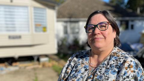 Phoenix Graham stands in the driveway of her home in Ruskington, Lincolnshire. She is wearing a floral blouse and dark-rimmed glasses. In the background stands a cream-coloured static caravan, next to a white-walled bungalow.