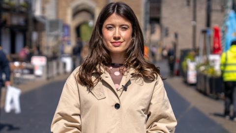 BBC News' Meleri Grug Williams, a young woman with long brown hair, looks down the camera with a shopping street and castle archway in the background.