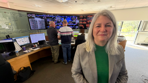 Sarah McManus, who is wearing a green jumper and a grey blazer, stands in front of the soon to be decommissioned system at the control room in South Gosforth. In the background are two controllers who are discussing how the network is currently operating.