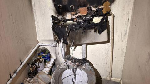 A burnt out toilet, with burnt wall, a burnt cistern and burnt items to the left. The room is full of soot. 