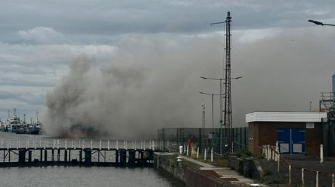 A large plume of grey smoke envelops a large cargo ship. It is visible behind a set of lock gates.