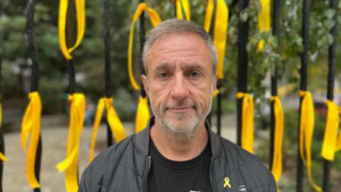 Ben Paul, wearing a small yellow ribbon, in front fo a black fence with yellow ribbons on