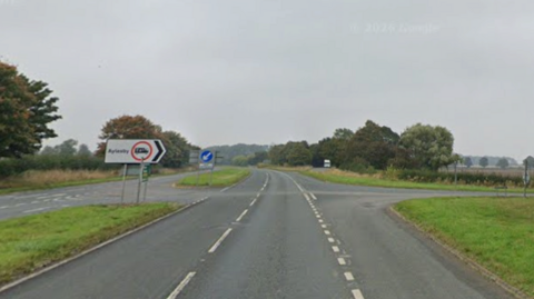 A dual carriageway junction. Grass verges can be seen on either side. The sky is grey.