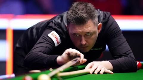 Kyren Wilson: A dark-haired man bending forward over a green snooker table aiming a light brown cue. He is wearing a long-sleeved black shirt which has the slogan "32 Red" written in red letters on a white patch at the top of the right sleeve. Two red snooker balls and a yellow snooker ball are sitting on the table.