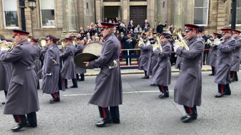 An Army band marches through Sunderland. Soldiers in uniform are playing trumpets and drums while marching past a crowd watching the procession.