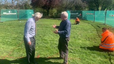 Two men with white hair talking on grass. There are metal fences with green tarp around them.