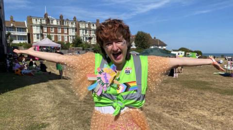 Amy Zing wearing a colourful outfit and a high-vis vest. She is smiling in the sun with her arms outstretched. Badges on her vest include a pride flag and a felt rosette that reads 'gayest town in Kent'.