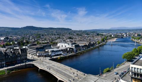 Ness Bridge shot from above on a sunny day, with views of Inverness city centre, the River Ness and hills in the background.