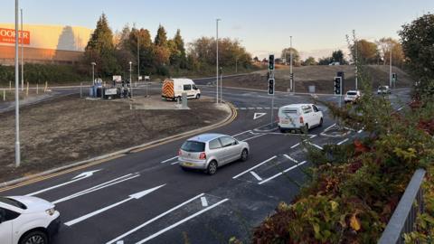 A freshly tarmacked road junction with crisp white road markings. The verges around the road are still covered with dirt and there is a van and some temporary traffic lights at the roadside. There are a couple of vehicles driving on the road.