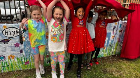 Five children named Jaylee, Mynah, Ashoka, Vishwan and Ananya stand in front of a mural they helped to paint. The mural is painted on a low wall that is topped with metal railings. The mural shows green grass at the bottom, a blue sky and buildings from Belfast's skyline. The children are holding up a red curtain that had been used to obscure the new mural before it was unveiled. 