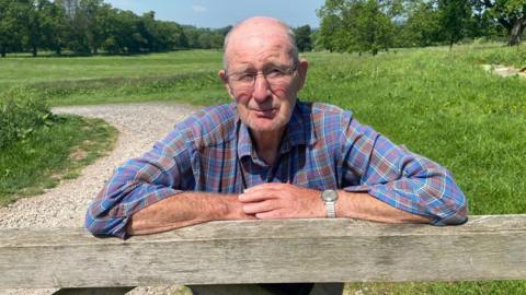 Ron Wood is a 78 year old man wearing a blue and brown shirt leaning on a wooden gate. there is a path and some fields behind with a number of trees on. He has thinning grey hair and glasses 