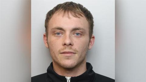 A man in a black zip up hoodie looks at the camera for his police mug shot.