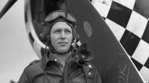 A black and white photo of Lt Lonnie Moseley wearing a fighter pilot's uniform. He has on a close-fitting hat, tied under his ears and goggles perched on the top of his head. Behind him is the propeller and nose of a fighter plane. 