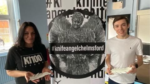 A man and a woman standing next to a poster for the Knife Angel while holding a white feather, which is a sneak preview of the statue due to be unveiled. 