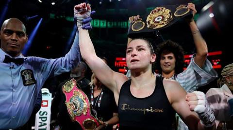 Katie Taylor, centre, with dark tied back hair, wearing a black vest with the word Champion on it. There is a man to her left wearing a blue shirt and a black bow tie, raising her right arm in the air.