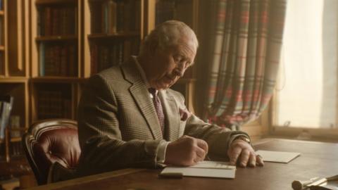 King Charles III writes a letter from a desk in front of a window in Buckingham Palace. He is wearing a blazer and a purple tie. 