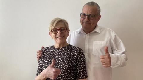 A smiling Val and Geoff King. She is wearing glasses and a black and white top. He is wearing a white shirt and both are holding their thumbs up in front of them. 