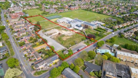 An aerial view of a school which has large playing fields either side. On the left, a playing field and car park is outlined with a red line. There are hundreds of homes around the school site.