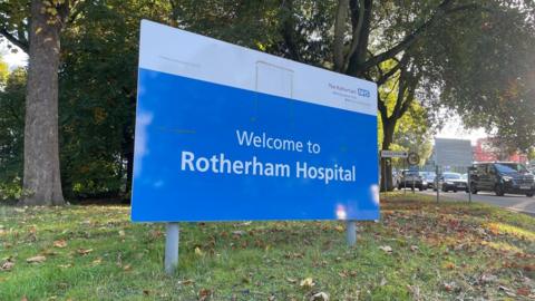 A large blue hospital sign on the grass outside a hospital with a large car park