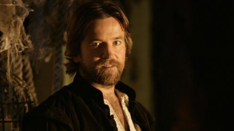 Head and shoulders shot of Dean Lennox Kelly playing Shakespeare in BBC drama Dr Who. He has a beard, a collar-less black jacket and white shirt and is in a dark room