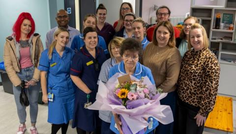 Cath Lyon in a nurse uniform holding a large bouquet of flowers. A group of colleagues are stood behind her. 