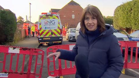 BBC reporter Sara Smith stood next to barriers. In the background are people in hi-viz clothing